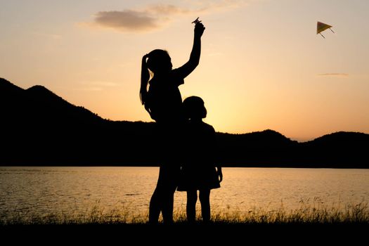 Silhouette of little daughter and mother playing a kite by the lake at sunset. Healthy summer activity for children. Funny time with family.
