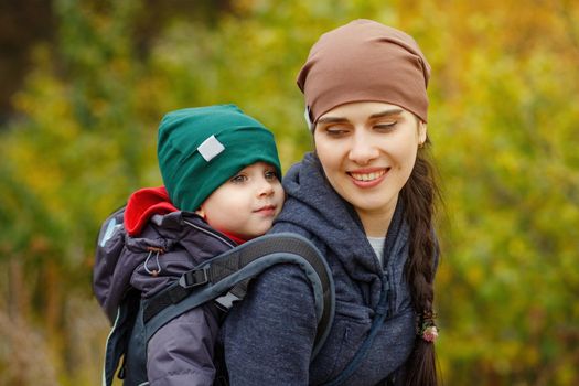 Young mother with her toddler kid boy on back in ergonomic baby carrier in autumn nature. Active mother concept.