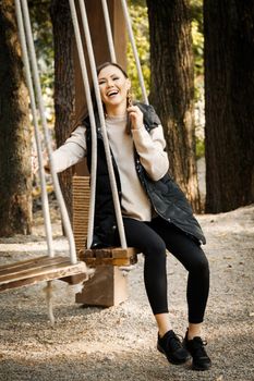 Happy young mongol woman with brown straight hair and makeup, riding a swing among outdoor autumn in warm clothes in basic colors in a park outdoors