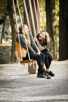 Vertical image of happy laughing couple sitting on swing in park outdoors.