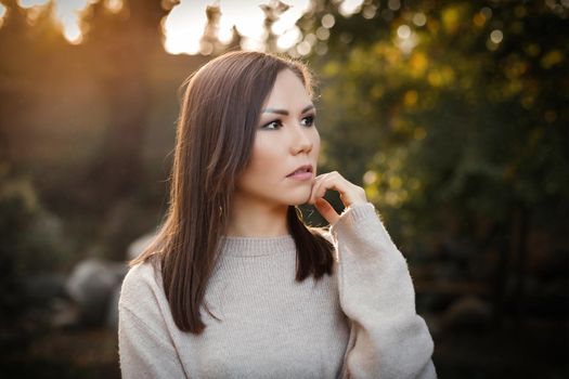 Pensive woman with hand near face in the park outdoor among trees at sunset. High quality photo