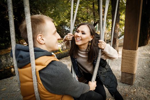 Cheerful young mixed race couple looking at each other while sitting on a swing outdoor.