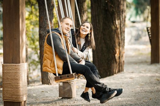 Portrait of young happy couple riding on a swing in autumn in warm clothes in basic colors in a park outdoors. High quality photo