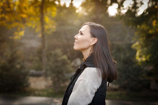 Profile of a young smiling woman with autumn in a park outdoors in a warm beige jacket and a black sleeveless jacket. High quality photo