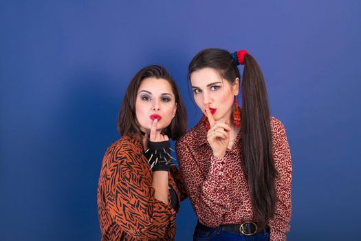 Two cute young women dressed in 90s style keeping secrets on purple background.