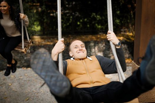 Young cheerful man swinging on a swing with legs raised high with his lover in the background in a forest park. High quality photo