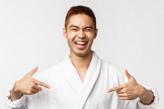 Beauty, spa and leisure concept. Portrait of successful, happy handsome asian man pointing fingers at himself and laughing, talking own accomplishment, wear bathrobe, white background.