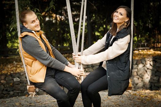 Cute young couple in the forest in the park sits on a swing and playfully looks at the camera. High quality photo