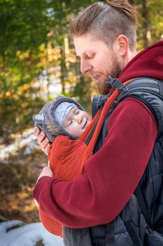 Side view of young bearded babywearing father with his baby in baby carrier walking outdoor.