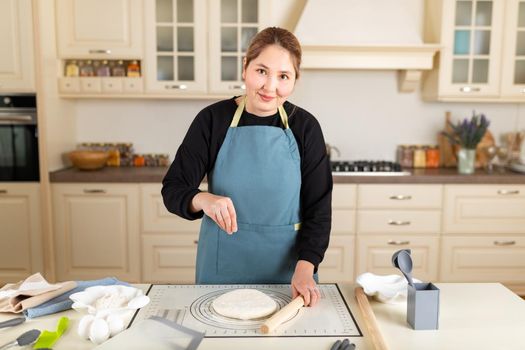 Asian woman cook in apron look at camera sprinkles flour on cooked wheat pastry dough on silicone baking mat in modern kitchen.