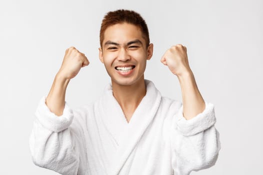 Beauty, spa and leisure concept. Satisfied asian man in bathrobe fist pump delighted, smiling broadly, looking pleased as winning prize, achieve goal, celebrating victory, white background.