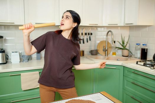 A young woman is fooling around and grimacing in the kitchen, singing into a rolling pin as if into a microphone.