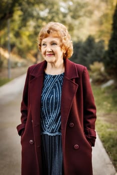 Smiling mature caucasian woman walking in autumn park outdoors CIS in light burgundy coat.
