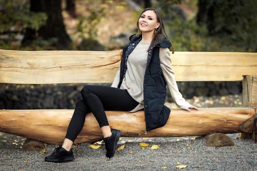 Portrait of a slender joyful young woman in full growth sitting on a wooden bench in a park outdoors. High quality photo