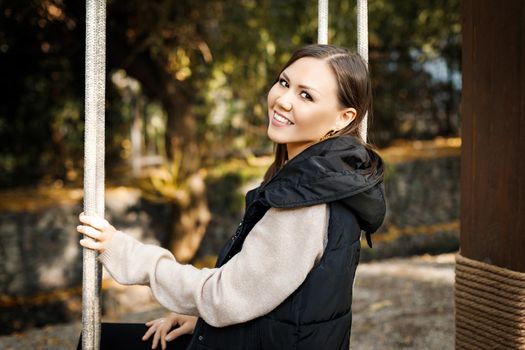 Beautiful young smiling woman sitting on a swing in autumn in a park outdoors. High quality photo