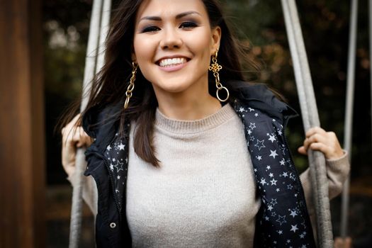 Portrait of a young happy asian woman with a snow-white smile swinging on a swing outdoor.