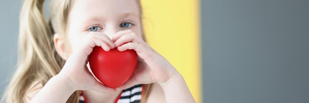 Portrait of little blonde girl holding plastic red heart, symbol for people, love, charity and family. Little girl needs mothers love. Mothers day, donation concept. Copy space in right