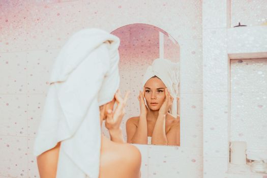 Young beautiful woman using face cream moisturizing lotion after bath. Pretty attractive girl in a towel on her head stands in front of a mirror in a home bathroom. Daily hygiene and skin care.