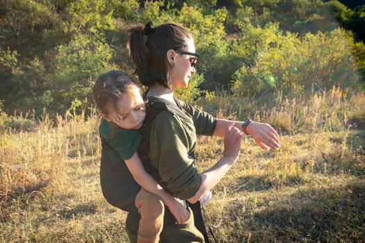 A woman looks at a fitness bracelet and uses an ergonomic baby back carrier for a child on a hiking trip.