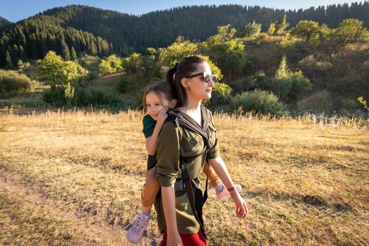 Young mother with her preschooler daughter on back in ergonomic baby carrier in autumn nature. Active mother concept.