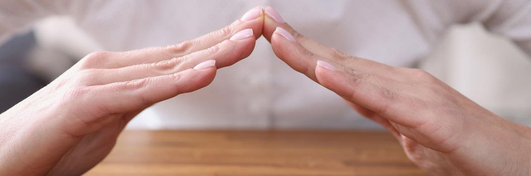 Close-up of female sitting at wooden table and forming hands house roof symbol with hands. Home safety and property security. Real estate insurance concept