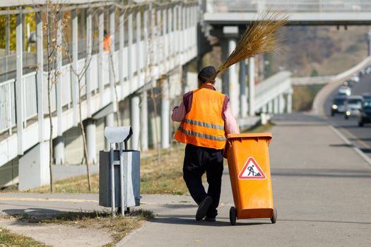City janitor in orange uniform with a broom and a mobile tank on wheels walks along the sidewalk. Almaty, Kazakhstan - 20 Oktober, 2021