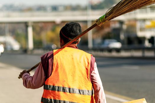 Back view of a city janitor in an orange uniform with a broom on his shoulder. Almaty, Kazakhstan - 20 Oktober, 2021