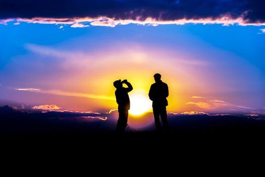 Two silhouettes of men talking and drinking a drink a jar at sunset or sunrise against an impressive sky and clouds.Outlines of people in the sunlight.Golden hour with friends on the horizon skyline