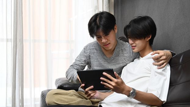 Cute gay couple using digital tablet on sofa, spending time at home. LGBT and love concept.