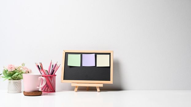 Black board with colorful sticky notes, pencil holder, coffee cup and flower pot on white table.
