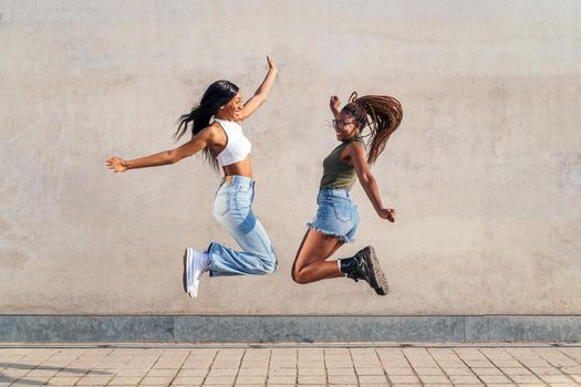 two young african girls jumping happy with a gray concrete wall in the background, concept of friendship and urban lifestyle