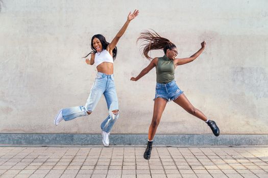 two young black girls jumping happy with a gray concrete wall in the background, concept of friendship and urban lifestyle