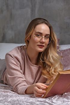 Blonde lady in glasses, casual clothing. She is looking at you, holding book, laying on bed in bedroom. Student, blogger, document studying. Interior with gray wall, pink blanket and pillows. Close-up