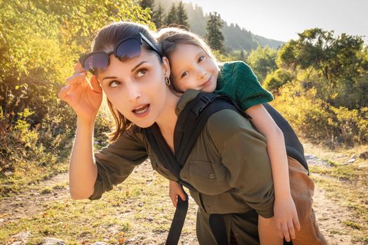 Happy preschool kid girl in babywearing sling at back her mother in hiking outdoor.