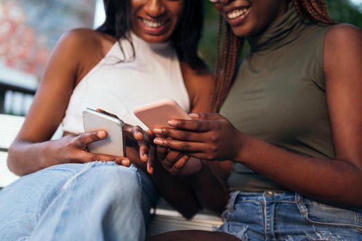 detail of the hands of two unrecognizable smiling black women looking at their mobile phones, concept of youth and communication