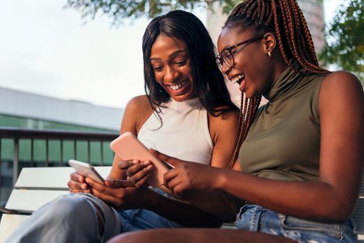 two young african women laughing happily while looking their mobile phones, concept of youth and communication, copy space for text