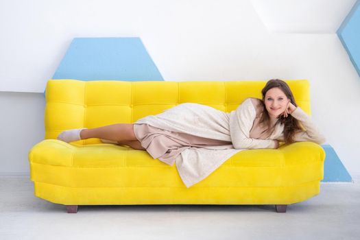 Young smiling caucasian woman 20 y.o. lies on a yellow couch, look at camera, in bright interior with copy space.