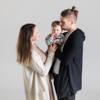 A man holds his child in his arms and kisses him while his wife looks at them on a white background.