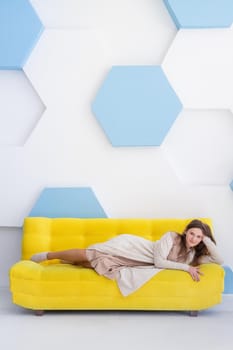 Young happy caucasian woman lies on a yellow sofa in bright interior with copy space, vertical.