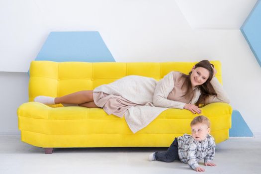 Young happy caucasian woman lies on a yellow sofa and watches her baby crawl on the floor. Waiting for baby's first steps.