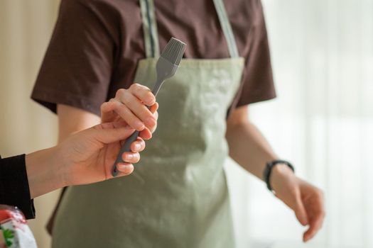 One cook passes a kitchen brush to another cook in a green apron.