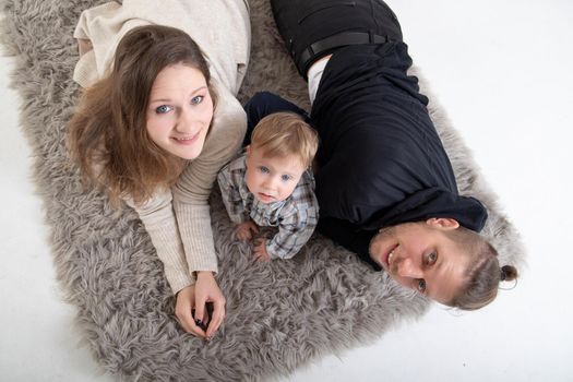 A young caucasian family with a child lies on a carpet on a white floor and looks at the camera. Top view.