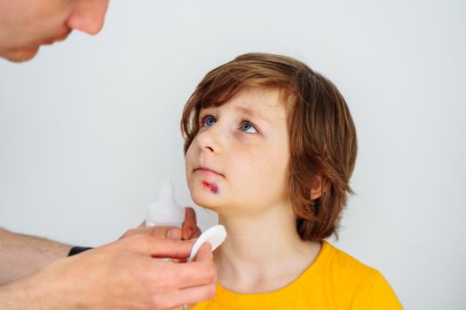 Dad doctor father treats bruised wound on his son school boy kid face. Man cleans addresses the sore wound on child face on white background with copy space for text.