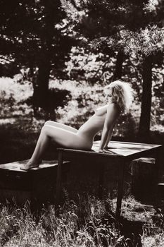 Fully naked beautiful young blonde woman relaxing in nature sitting at a picnic table among pine trees. Retro style imitation.