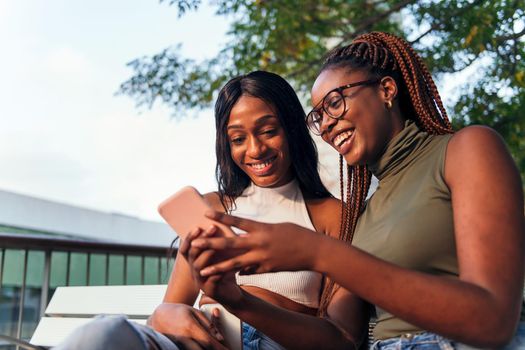 two young african women having fun sitting on a park bench with a mobile phone, concept of youth and communication, copy space for text