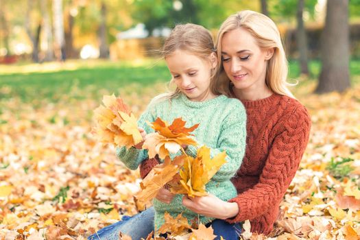 Happy young caucasian woman and little girl holding autumn yellow leaves sitting at the park