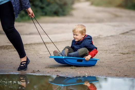 Cute boy playing with sled in summertime. Mother towing son on sleigh, portrait.