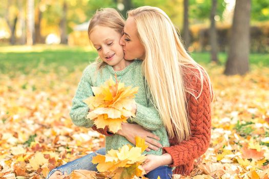 Happy young caucasian mother and little daughter holding autumn yellow leaves sitting and kissing at the park