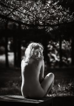 Fully naked beautiful young blonde woman relaxing in nature sitting at a picnic table among pine trees. Black and white image