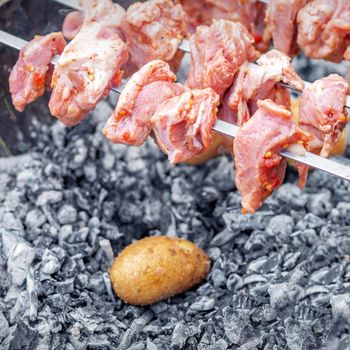 Hands of man prepares barbecue meat with potatoes on skewer by grill on fire outdoors. Concept of lifestyle rustic food preparation